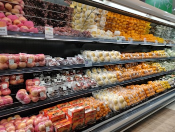 A supermarket produce section displays a neatly organized assortment of fruits. The shelves are filled with apples, grapes, pears, oranges, and other fruits, each packed in individual wrapping or foam nets. Pricing labels are visible above the fruits, indicating price per kilogram.