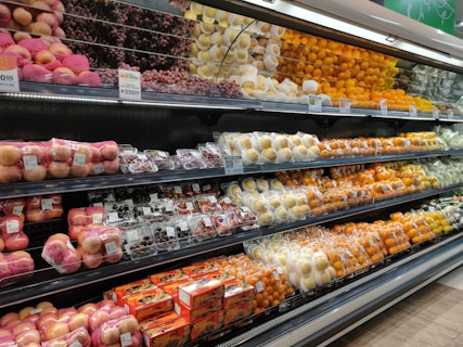 A supermarket produce section displays a neatly organized assortment of fruits. The shelves are filled with apples, grapes, pears, oranges, and other fruits, each packed in individual wrapping or foam nets. Pricing labels are visible above the fruits, indicating price per kilogram.