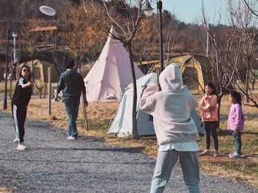 A group of people playing frisbee in a park-like setting with tents in the background. One person is actively throwing the frisbee while another is preparing to catch it. Two children are watching nearby, standing on a gravel path surrounded by trees and grass.