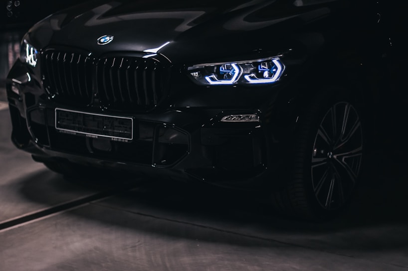 A sleek black car is parked in a dimly lit environment. The focus is on the front end, highlighting its modern, luminous headlights and robust grill design.
