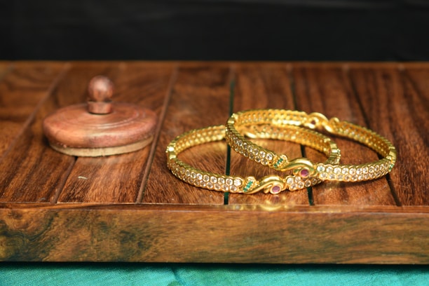 On a wooden surface, two ornamental gold bangles are adorned with embedded jewels, featuring sparkling, multicolored stones. A wooden container with a lid sits nearby, adding a rustic touch to the elegant display.