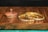 On a wooden surface, two ornamental gold bangles are adorned with embedded jewels, featuring sparkling, multicolored stones. A wooden container with a lid sits nearby, adding a rustic touch to the elegant display.