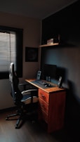 A compact workspace featuring a wooden desk with three drawers, an ergonomic office chair, and a computer setup with a monitor, keyboard, mouse, and speakers. The room is dimly lit, with a window covered by blinds letting in minimal light. A shelf above the desk holds several books and a small decorative item.