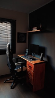 A compact workspace featuring a wooden desk with three drawers, an ergonomic office chair, and a computer setup with a monitor, keyboard, mouse, and speakers. The room is dimly lit, with a window covered by blinds letting in minimal light. A shelf above the desk holds several books and a small decorative item.