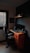 A compact workspace featuring a wooden desk with three drawers, an ergonomic office chair, and a computer setup with a monitor, keyboard, mouse, and speakers. The room is dimly lit, with a window covered by blinds letting in minimal light. A shelf above the desk holds several books and a small decorative item.