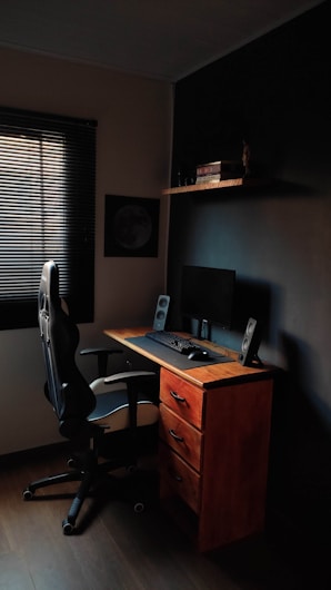 A compact workspace featuring a wooden desk with three drawers, an ergonomic office chair, and a computer setup with a monitor, keyboard, mouse, and speakers. The room is dimly lit, with a window covered by blinds letting in minimal light. A shelf above the desk holds several books and a small decorative item.