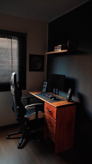 A compact workspace featuring a wooden desk with three drawers, an ergonomic office chair, and a computer setup with a monitor, keyboard, mouse, and speakers. The room is dimly lit, with a window covered by blinds letting in minimal light. A shelf above the desk holds several books and a small decorative item.