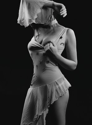 A monochrome image featuring a woman partially obscured by a flowing piece of fabric. She is wearing a sleeveless dress with an asymmetrical hem and geometric designs. The pose is expressive, with one arm raised, creating dramatic shadows and highlights.