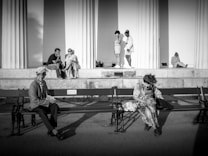 A black and white photograph depicting people in a public space. In the foreground, two individuals sit on separate benches, each occupied with their own thoughts. In the background, five more individuals are near large columns, engaged in various activities such as sitting, standing, and conversing. The strong shadows and architectural elements create a dramatic atmosphere.