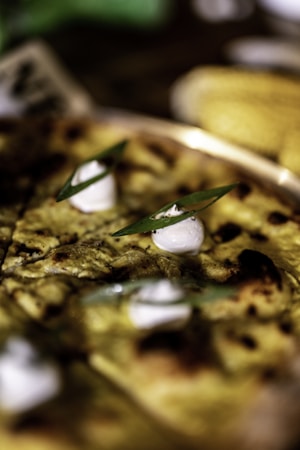 A close-up of a savory dish with a golden-brown crust, garnished with dollops of white cream and green herbs. The texture appears crispy with charred spots, suggesting it might be freshly baked. The background is softly blurred, focusing attention on the dish.