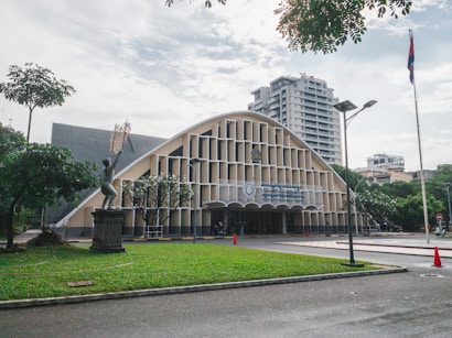 An architectural building with a unique curved roof and vertical slats on the facade. In the foreground, there is a statue of a person holding up an object, surrounded by greenery and pathways. The background features a modern high-rise building and a flagpole with a flag. The area is well-maintained, with trees and manicured grass, and some traffic cones are visible near the entrance.