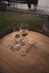 Two glasses of white wine are placed on a wooden barrel, which is engraved with the words 'Bodegas Navajas' along with some decorative elements and additional text. The scene is set outdoors, and the background appears slightly blurred, suggesting a relaxed, picturesque environment.