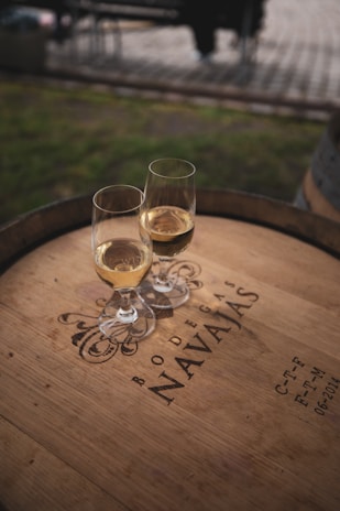 Two glasses of white wine are placed on a wooden barrel, which is engraved with the words 'Bodegas Navajas' along with some decorative elements and additional text. The scene is set outdoors, and the background appears slightly blurred, suggesting a relaxed, picturesque environment.