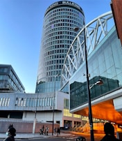 A modern urban scene featuring a tall cylindrical skyscraper with the label 'BULLRING' at the top. The building has a glass facade with a grid pattern. Surrounding it are other contemporary structures with reflective surfaces, creating a complex architectural composition. The scene is populated with people walking along the street, and there's a bridge or overpass made of white structural elements.