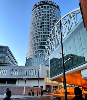 A modern urban scene featuring a tall cylindrical skyscraper with the label 'BULLRING' at the top. The building has a glass facade with a grid pattern. Surrounding it are other contemporary structures with reflective surfaces, creating a complex architectural composition. The scene is populated with people walking along the street, and there's a bridge or overpass made of white structural elements.