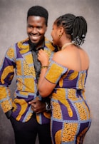 Two people are smiling and engaging with each other while dressed in matching colorful, patterned outfits. The background is a neutral gray, focusing attention on the individuals and their attire.