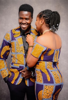 Two people are smiling and engaging with each other while dressed in matching colorful, patterned outfits. The background is a neutral gray, focusing attention on the individuals and their attire.