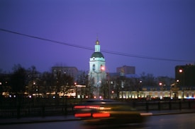 A brightly illuminated church with a green dome stands against a twilight sky. In the foreground, blurred lights from passing vehicles create streaks of red and orange. Buildings and bare trees surround the church, with scattered streetlights adding a warm glow to the scene.