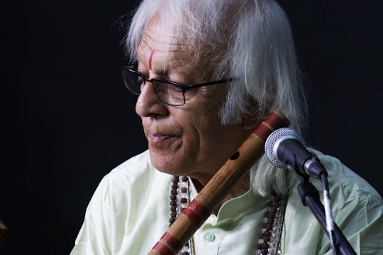 An elderly man with white hair and glasses is holding a traditional wooden flute near his shoulder. He is wearing a light-colored shirt and a beaded necklace. A microphone is positioned in front of him, suggesting a musical performance or recording setting. The background is dark, which highlights the subject.