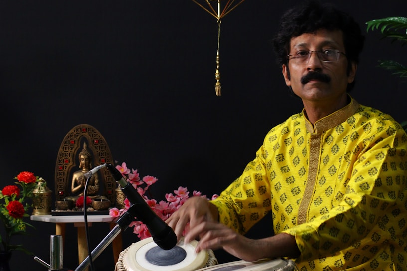 A man in a bright yellow patterned shirt is playing a tabla. He is sitting beside a decorative setup, including a bronze statue of a seated figure on a table adorned with red and pink flowers. A microphone is positioned near the tabla.
