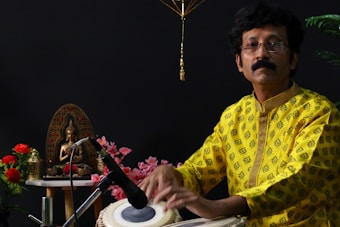 A man in a bright yellow patterned shirt is playing a tabla. He is sitting beside a decorative setup, including a bronze statue of a seated figure on a table adorned with red and pink flowers. A microphone is positioned near the tabla.