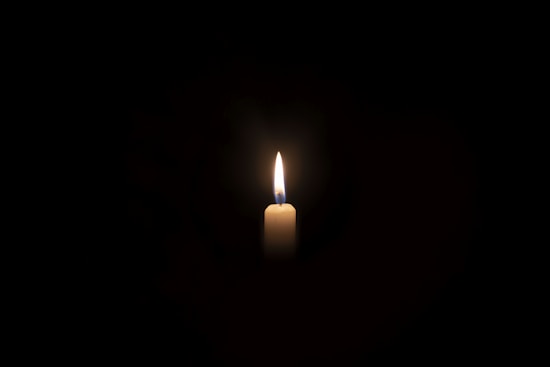 A single, lit white candle stands in the center against a dark background. The flame flickers gently, casting a soft glow and subtle shadows.