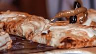 A close-up of a cheesy, baked dish topped with a green chili and an olive, possibly a type of calzone or stuffed bread. The dish appears to be served on a patterned paper, showcasing gooey melted cheese with a golden-brown crust.