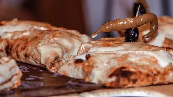 A close-up of a cheesy, baked dish topped with a green chili and an olive, possibly a type of calzone or stuffed bread. The dish appears to be served on a patterned paper, showcasing gooey melted cheese with a golden-brown crust.
