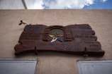 A wooden sign mounted on a textured wall, featuring inscriptions in multiple languages including English, Hebrew, and Arabic. The text mentions the 'Carmel Osfiye heritage center and the development of the Druze woman.' At the center of the sign is a stylized arch and stars in green, red, blue, yellow, and white. Contact numbers are displayed at the bottom.