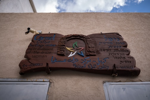 A wooden sign mounted on a textured wall, featuring inscriptions in multiple languages including English, Hebrew, and Arabic. The text mentions the 'Carmel Osfiye heritage center and the development of the Druze woman.' At the center of the sign is a stylized arch and stars in green, red, blue, yellow, and white. Contact numbers are displayed at the bottom.