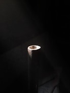 A cylindrical metallic pipe stands vertically against a dark background. Light casts a soft glow on the top surface, highlighting its rough, textured surface and casting subtle shadows.