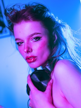 A person with bright makeup and styled hair holds a pair of headphones against a vibrant, neon-lit background. The lighting casts a mix of blue and pink hues, highlighting the individual's facial features and expression.