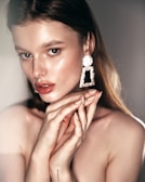 A woman with straight, brown hair gazes intently. She wears large, ornate earrings and has a subtle glow on her skin. Her hands are raised near her face with neatly manicured nails.