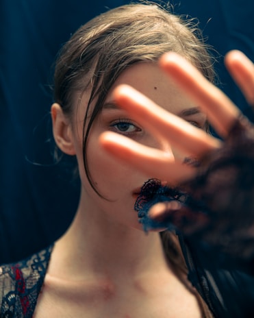 A person partially obscured by a hand held in front of their face, with the focus on their eye visible through the fingers. The lighting creates a soft, intimate atmosphere, highlighting their features. Lace fabric and a dark background add an element of mystery.