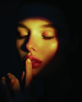 A woman's hand gently touching her lips, highlighted by a single beam of warm amber light in a dark room.