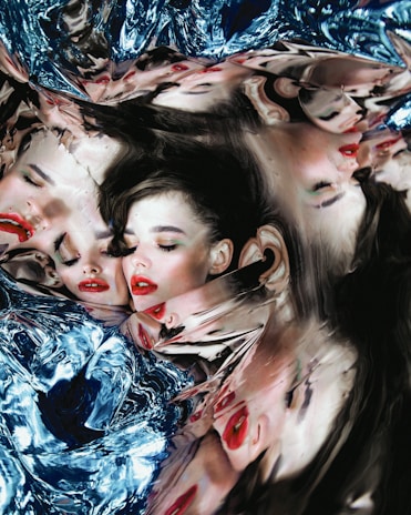Multiple distorted reflections of a woman's face appear in a warped and fluid-like surface, creating an abstract and surreal visual effect. The mirrored images blend seamlessly with a metallic blue texture that resembles liquid or melted glass.