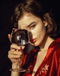 A woman holds a glass of water up to her face, partially obscuring her features. Her hair is loosely styled, and she is wearing a red garment with floral embroidery. The lighting creates deep shadows and highlights across her face, adding a dramatic and introspective quality to the image.