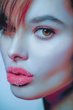 A close-up artistic portrait of a person with vivid pink lips coated in sugar. The subject's skin has a bluish hue, and their eyes are emphasized with dramatic makeup. Hair partially covers one side of the face, adding a mysterious element.