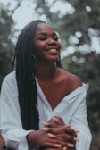 A person with long braided hair and hoop earrings is smiling warmly, wearing a white off-the-shoulder shirt. The background is blurred with soft green tones, suggesting an outdoor setting.