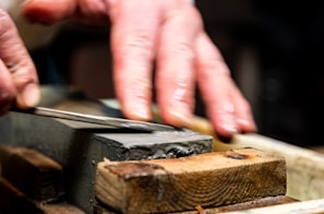 A pair of hands sharpening a knife on a whetstone, with a close-up focus on the blade and stone. The hands appear skillful, applying careful pressure for precise sharpening.