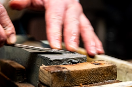 A pair of hands sharpening a knife on a whetstone, with a close-up focus on the blade and stone. The hands appear skillful, applying careful pressure for precise sharpening.