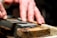 A pair of hands sharpening a knife on a whetstone, with a close-up focus on the blade and stone. The hands appear skillful, applying careful pressure for precise sharpening.