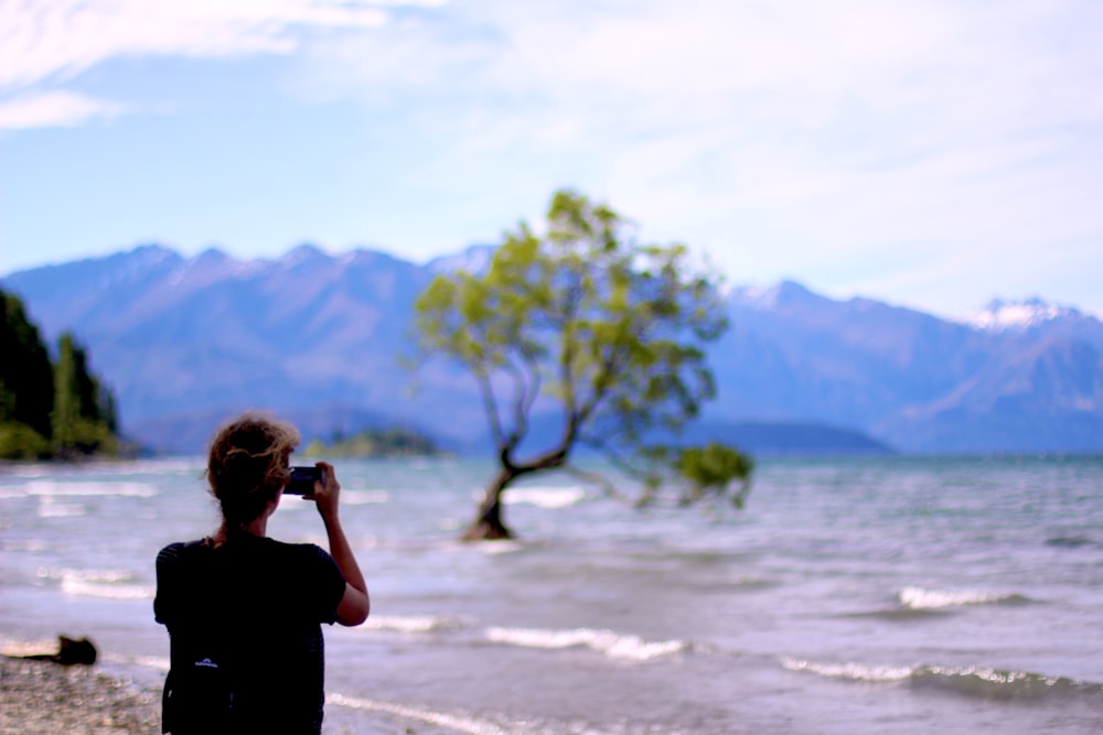 a woman taking a picture of a tree by the water