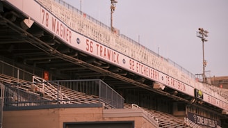 A view of an empty sports stadium featuring metal bleachers and a signage displaying numbers and names like Wagner, Trawick, Etcheverry, and Patterson. Floodlights are visible in the background, and the atmosphere suggests a calm and quiet moment.