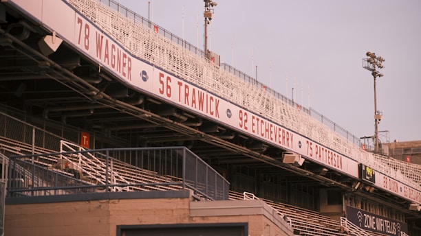 A view of an empty sports stadium featuring metal bleachers and a signage displaying numbers and names like Wagner, Trawick, Etcheverry, and Patterson. Floodlights are visible in the background, and the atmosphere suggests a calm and quiet moment.