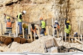 Workers wearing safety gear are engaged in construction activities at a site with rocky terrain. They are handling large metal rods, possibly for reinforcing concrete structures. Piles of earth and construction materials surround the area.