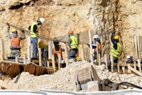 Workers wearing safety gear are engaged in construction activities at a site with rocky terrain. They are handling large metal rods, possibly for reinforcing concrete structures. Piles of earth and construction materials surround the area.