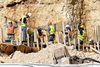 Workers wearing safety gear are engaged in construction activities at a site with rocky terrain. They are handling large metal rods, possibly for reinforcing concrete structures. Piles of earth and construction materials surround the area.