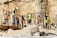 Workers wearing safety gear are engaged in construction activities at a site with rocky terrain. They are handling large metal rods, possibly for reinforcing concrete structures. Piles of earth and construction materials surround the area.