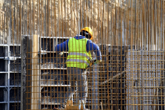 A construction worker wearing a yellow safety vest, blue sweater, and yellow hard hat is seen working among steel bars. The worker appears to be assembling or inspecting a structure made of metal and concrete.